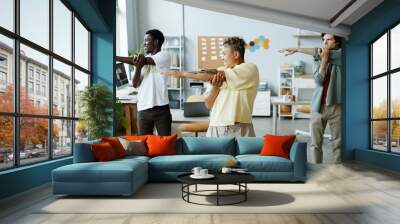 Diverse group of smiling young people doing stretching exercise during warm up break in office Wall mural