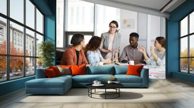 Diverse group of young business people discussing project sitting at table in modern office, focus on female manager giving instructions Wall mural