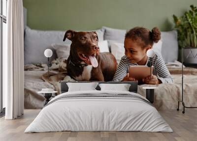 Front view portrait of cute African-American girl lying on bed with big pet dog and smiling, copy space Wall mural