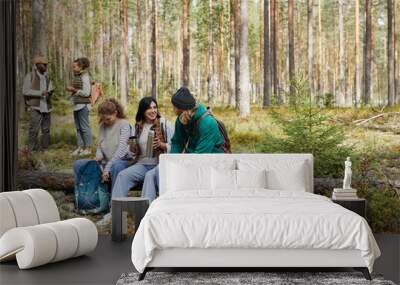 Full length portrait of diverse group of friends taking break while enjoying hike in forest, focus on three people sitting on log, copy space Wall mural