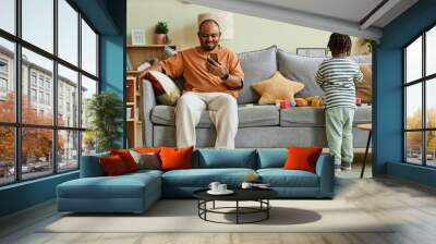 Full length portrait of father using smartphone with son playing on sofa at home, copy space Wall mural