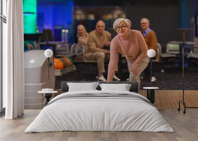 Full length portrait of joyful senior woman playing bowling with group of friends in background, copy space Wall mural