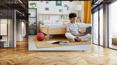 Full length portrait of mixed-race teenage boy sitting on floor in cozy room and using laptop, copy space Wall mural