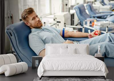 Full length portrait of young man donating blood while lying in comfortable chair at med center and looking at camera Wall mural