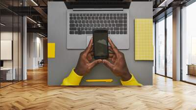 Graphic gray and yellow background of African-American male hands holding smartphone with blank screen over laptop while working at desk, top down view, copy space Wall mural