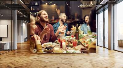 Group of emotional young people enjoying dinner party with friends and smiling happily sitting at table in dimly lit room, copy space Wall mural