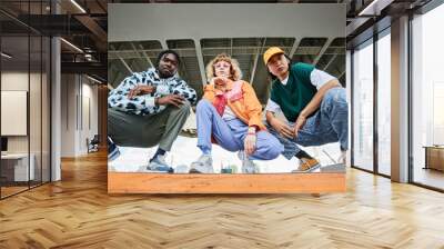 Group of three young people wearing street style clothes outdoors while sitting on stairs in urban area and looking at camera Wall mural