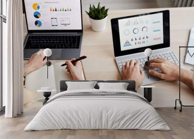 Hands of two young coworkers sitting in front of laptop screens with charts and diagrams and analyzing graphic data by workplace Wall mural