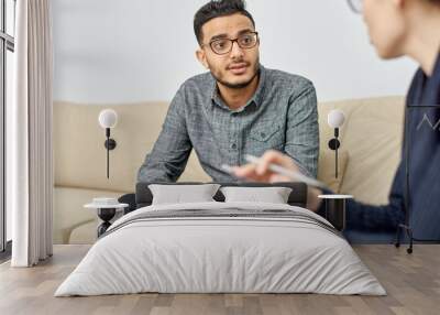 Handsome young man suffering from depression sitting on cozy sofa and listening to highly professional psychologist while she giving him piece of advice Wall mural