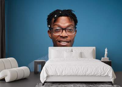 Head and shoulders portrait of young African-American man smiling at camera while posing against blue background, copy space Wall mural