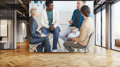 High angle view at multi-ethnic group of people sitting on chairs in support group circle and sharing stories with psychologist Wall mural