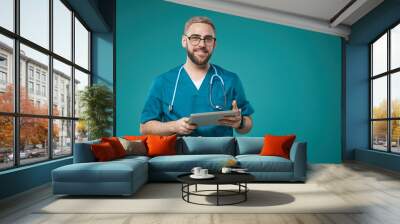 Horizontal medium studio portrait of Caucasian medical worker wearing blue uniform holding tablet computer looking at camera smiling Wall mural