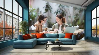 Jolly confident enterprising ladies in casual shirts sitting at table in modern cafe and viewing papers while working on report Wall mural