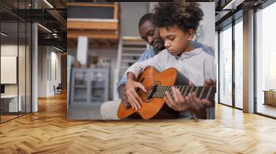 Mature man spending spare time with his son at home, teaching him how to play beautiful song on guitar Wall mural