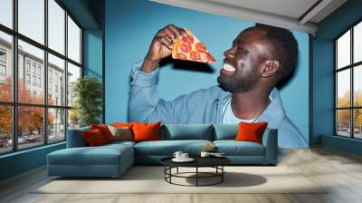 Medium close up shot of smiling young Black man standing with eyes closed while holding appetizing slice of pepperoni pizza near face, copy space Wall mural