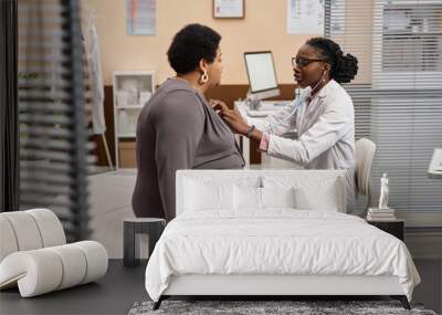 Medium long shot of young Black woman doctor using stethoscope listening to mature female patient respiration while sitting in hospital room Wall mural