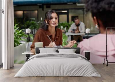 Medium shot of businesswoman talking to female colleague at business meeting discussing work strategy while sitting at cafe table in contemporary office center, copy space Wall mural