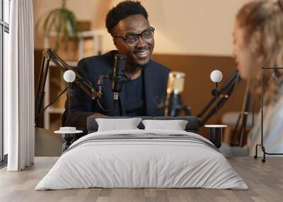 Medium shot of cheerful African American man wearing glasses and black suit talking to unrecognizable female guest sitting at table during podcast in studio Wall mural