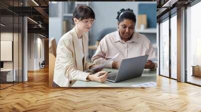 Medium shot of two smiling diverse female coworkers holding papers looking at laptop while sitting at desk in office Wall mural