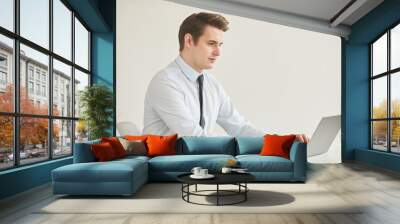 Minimal side view portrait of successful young businessman using laptop while sitting at desk and working in office against white wall, copy space Wall mural