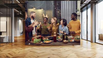 Multi-ethnic group of smiling young people enjoying dinner together standing at table in modern interior and holding wine glasses, copy space Wall mural