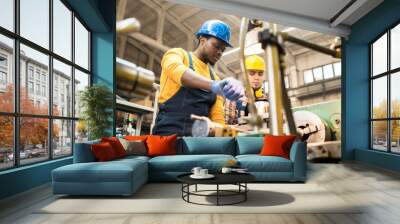 Multi-ethnic team of workers wearing overalls and protective helmets using lathe in order to machine workpiece, interior of spacious production department on background Wall mural