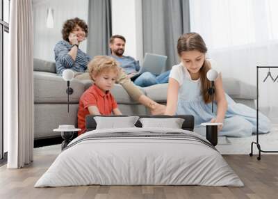 Ordinary day of large family: curly little boy and his elder sister sitting on carpet and drawing pictures with pencils while their pretty mother talking on phone and their bearded dad using laptop Wall mural