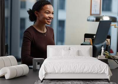 Portrait of black young woman using computer at desk in office and smiling enjoying work Wall mural