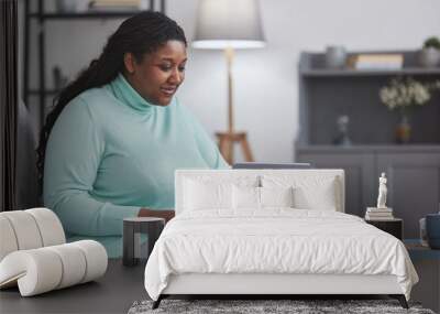 Portrait of curvy African American woman using laptop and smiling while enjoying work from home sitting on couch in minimal grey interior, copy space Wall mural