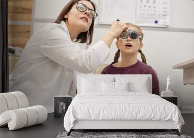 Portrait of cute teenage girl wearing trial frame during childs vision test in modern ophthalmology clinic Wall mural