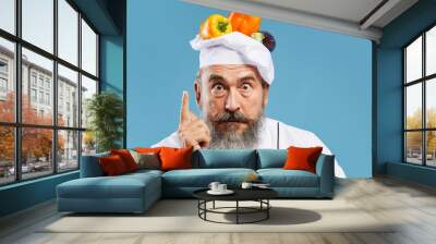 Portrait of excited bearded chef looking at camera and pointing up while posing against blue background with fresh vegetables on top of hat Wall mural