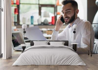 Portrait of handsome bearded businessman wearing white shirt speaking by phone and reading documents while working at table in office or cafe Wall mural