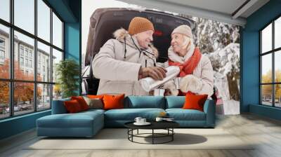 Portrait of happy mature couple enjoying cup of hot coco outdoors in winter while sitting in car trunk Wall mural