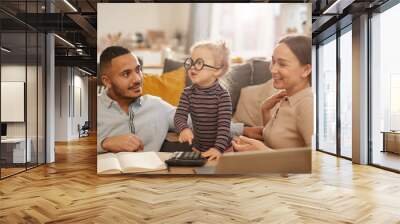 Portrait of happy young family counting home finances with cute little girl wearing glasses in sunlit apartment Wall mural