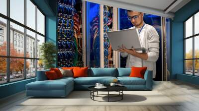 Portrait of modern young man holding laptop standing in server room working with supercomputer in blue light Wall mural