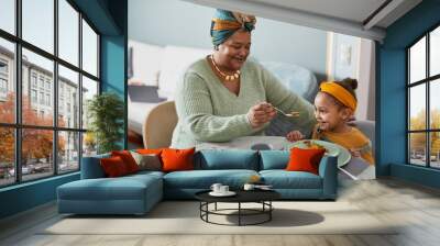 Portrait of smiling African-American grandma feeding cute little girl while enjoying dinner with family in home interior, copy space Wall mural