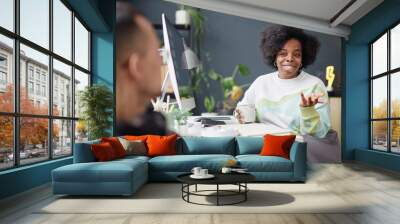 Portrait of smiling African American woman talking to colleague while sitting at desk in modern office and taking break copy space Wall mural