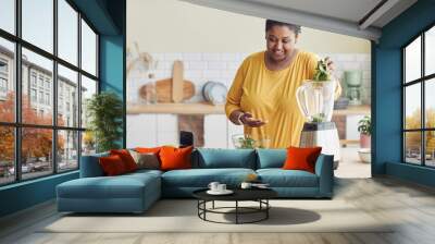 Portrait of smiling black woman using blender while making healthy meal in kitchen and filming cooking video, copy space Wall mural