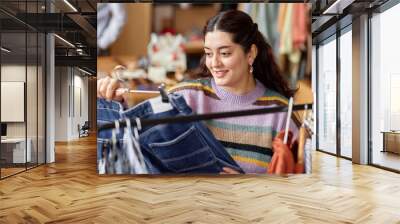 Portrait of smiling Middle Eastern female customer holding hanger with second hand pants choosing vintage gently used clothes at thrift store, copy space Wall mural