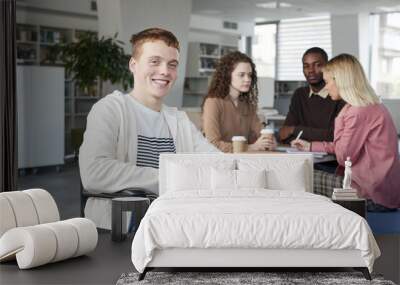 Portrait of smiling red haired boy using wheelchair studying with group of students in college library and looking at camera, copy space Wall mural