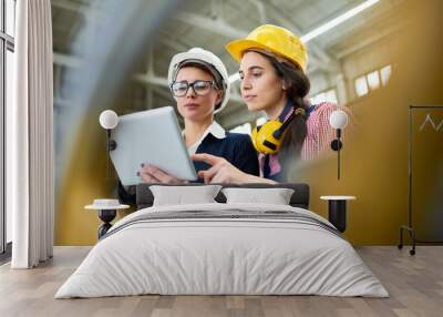 Portrait of two female engineers wearing hardhats standing in spacious workshop of modern plant using digital tablet Wall mural