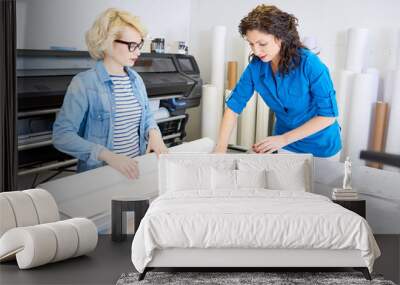 Portrait of two modern young women working in printing shop or publishing company, loading machines with paper and cutting brochures Wall mural