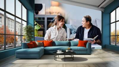 Portrait of two smiling modern women using laptops and communicating while working from home together Wall mural