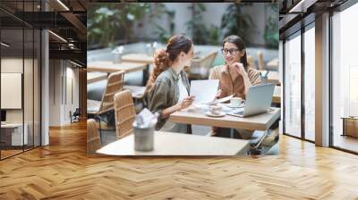 Portrait of two young businesswomen discussing project while sitting at table in designer cafe, copy space Wall mural