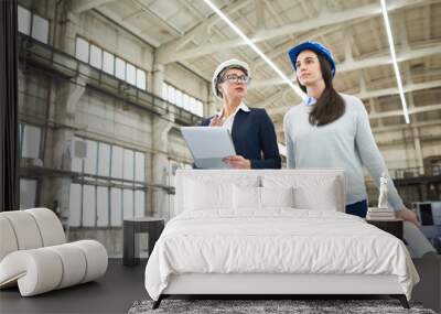 Portrait of two young female engineers wearing hardhats discussing work while crossing  workshop of modern factory holding plans , copy space Wall mural