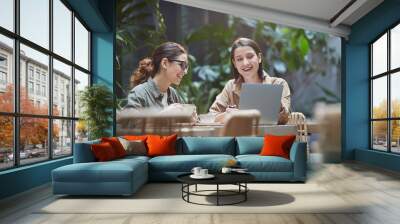 Portrait of two young women laughing happily while using laptop on outdoor cafe terrace decorated with plants, copy space Wall mural