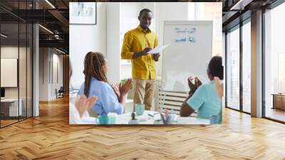 Portrait of young African-American man standing by whiteboard in conference room and smiling with multi-ethnic business team applauding, copy space Wall mural