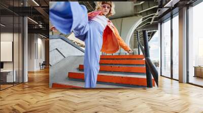 Portrait of young carefree woman wearing street style fashion outdoors in urban setting and kicking up to camera Wall mural