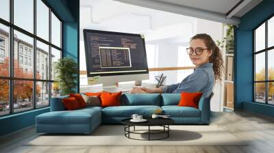 Portrait of young woman looking at camera while writing programming code on computer screen and working at desk in contemporary office interior, copy space Wall mural