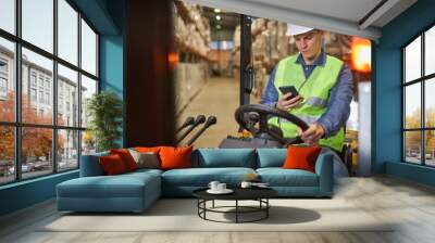 Portrait of young worker driving forklift truck in warehouse and using smartphone Wall mural